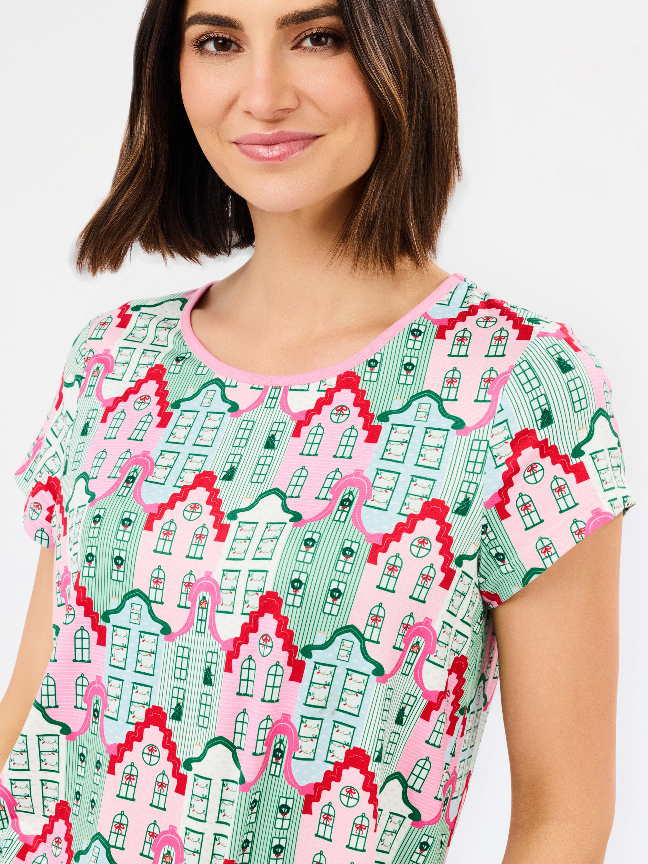 A woman wearing a short-sleeve crewneck top with a colorful repeating pattern of stylized houses in pink, green, and red on a light background.