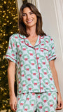 A woman wearing a matching holiday-themed pajama set with a light blue background, green wreath pattern, and red bow details, standing in front of a decorated Christmas tree.
