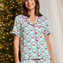A woman wearing a matching holiday-themed pajama set with a light blue background, green wreath pattern, and red bow details, standing in front of a decorated Christmas tree.