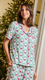 A woman wearing a matching short-sleeve pajama set with a green wreath and red bow holiday print, standing indoors in front of a lit Christmas tree.