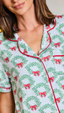 A close-up of a woman wearing holiday-themed short-sleeve pajamas with a green wreath and red bow pattern, red piping, and a white base, set against a softly blurred background with warm lights.