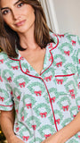 A woman wearing a short-sleeve pajama top with a green wreath and red bow holiday print, featuring red piping trim, posed in front of a softly blurred festive background.