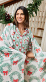 A woman wearing a matching holiday-themed pajama set and wrapped in a plush throw blanket, standing on indoor stairs decorated with Christmas garlands.