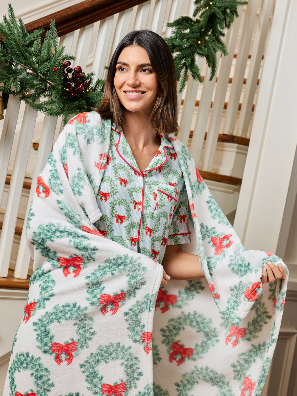 A woman wearing a matching holiday-themed pajama set and wrapped in a plush throw blanket, standing on indoor stairs decorated with Christmas garlands.