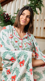A woman wearing a white holiday pajama set with green wreaths and red bows, wrapped in a matching throw blanket, standing in front of a staircase decorated with a green garland.