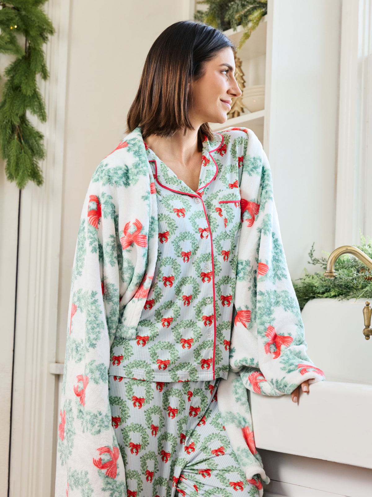 A woman wearing a matching holiday-themed pajama set with a white base, green wreath pattern, and red bow accents, standing in a bright indoor setting next to a sink.