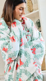 A woman wrapped in a soft white blanket featuring a green toile-style floral pattern and coral-red bow motifs, standing in a bright indoor room.