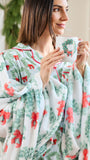 A woman wearing a soft white holiday-themed blanket with red bows and green wreath patterns, holding a white mug with a green wreath design. The background is a softly blurred indoor setting with warm lighting.