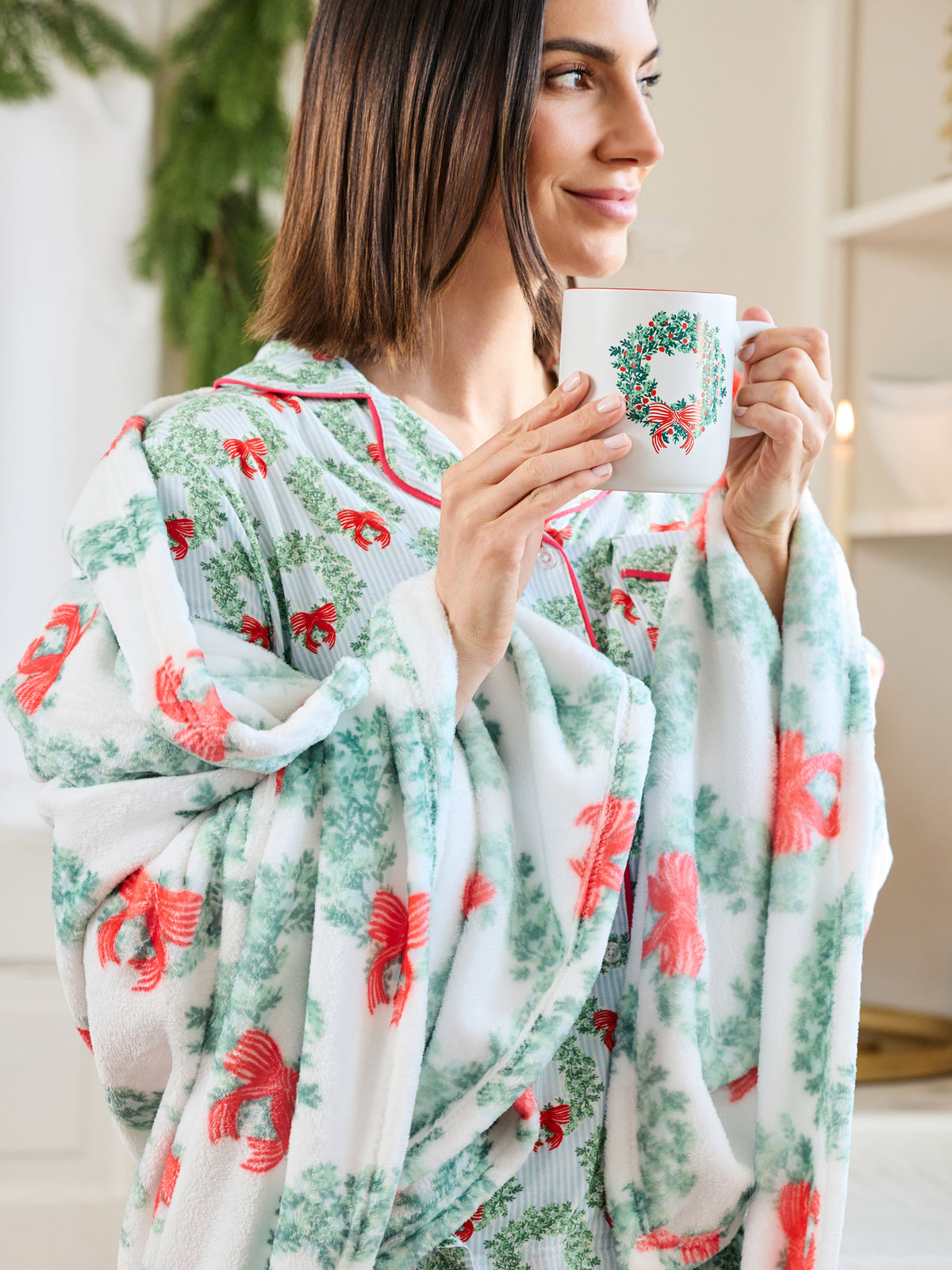 A woman wearing a soft white holiday-themed blanket with red bows and green wreath patterns, holding a white mug with a green wreath design. The background is a softly blurred indoor setting with warm lighting.