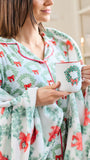 A woman wearing a festive holiday-themed pajama set with a matching throw blanket. The set features a white base with a green holly wreath pattern and red bows, accented by red piping.