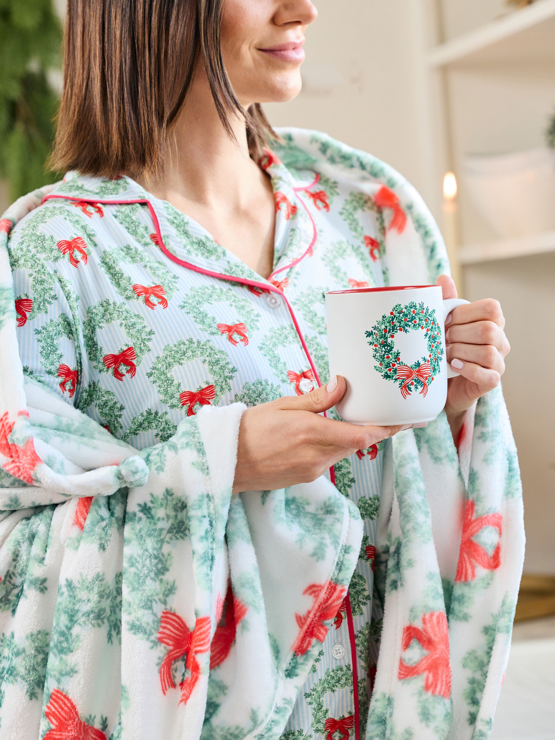 A woman wearing a festive holiday-themed pajama set with a matching throw blanket. The set features a white base with a green holly wreath pattern and red bows, accented by red piping.