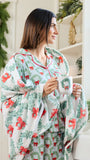 A woman wearing a festive Christmas-themed pajama set with a matching throw blanket, holding a mug decorated with a wreath. The pajamas and blanket feature a white base with green pine garland patterns and red bows, set in a bright, holiday-decorated room.