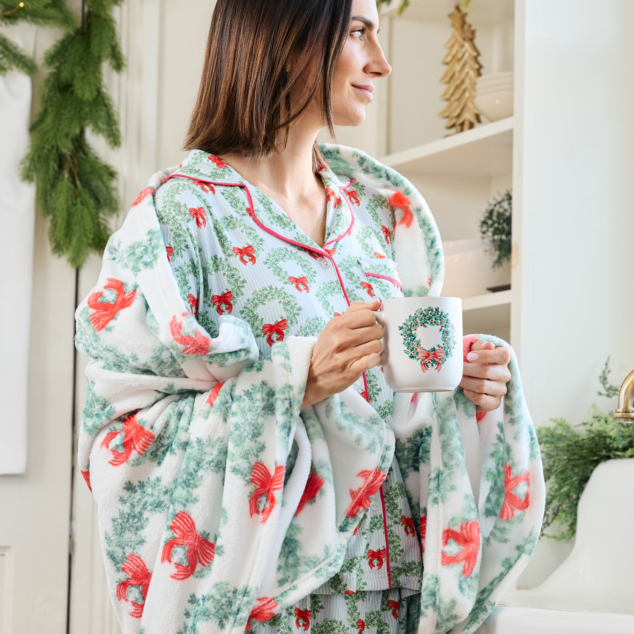 A woman wearing a festive Christmas-themed pajama set with a matching throw blanket, holding a mug decorated with a wreath. The pajamas and blanket feature a white base with green pine garland patterns and red bows, set in a bright, holiday-decorated room.