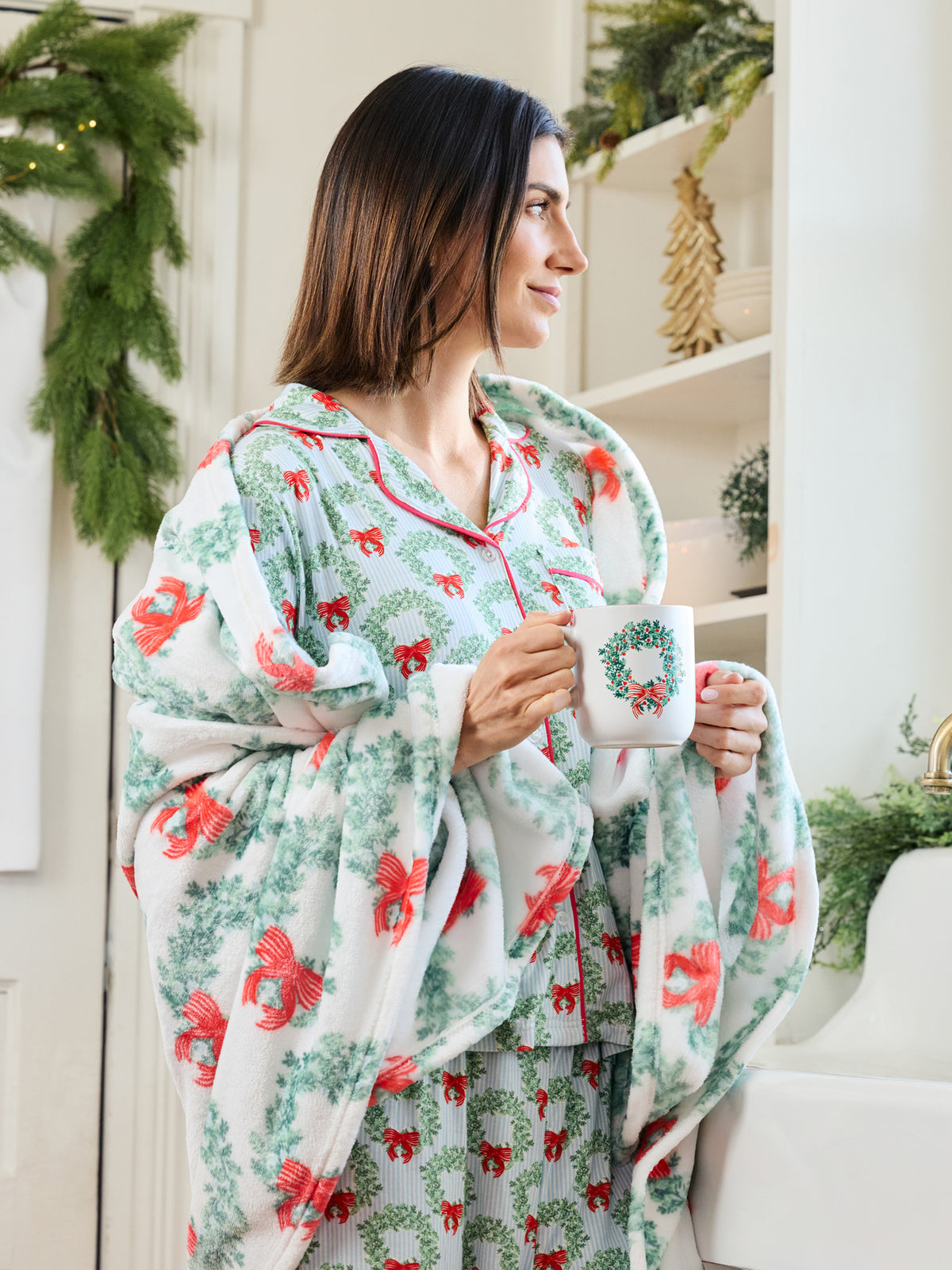A woman wearing a festive Christmas-themed pajama set with a matching throw blanket, holding a mug decorated with a wreath. The pajamas and blanket feature a white base with green pine garland patterns and red bows, set in a bright, holiday-decorated room.