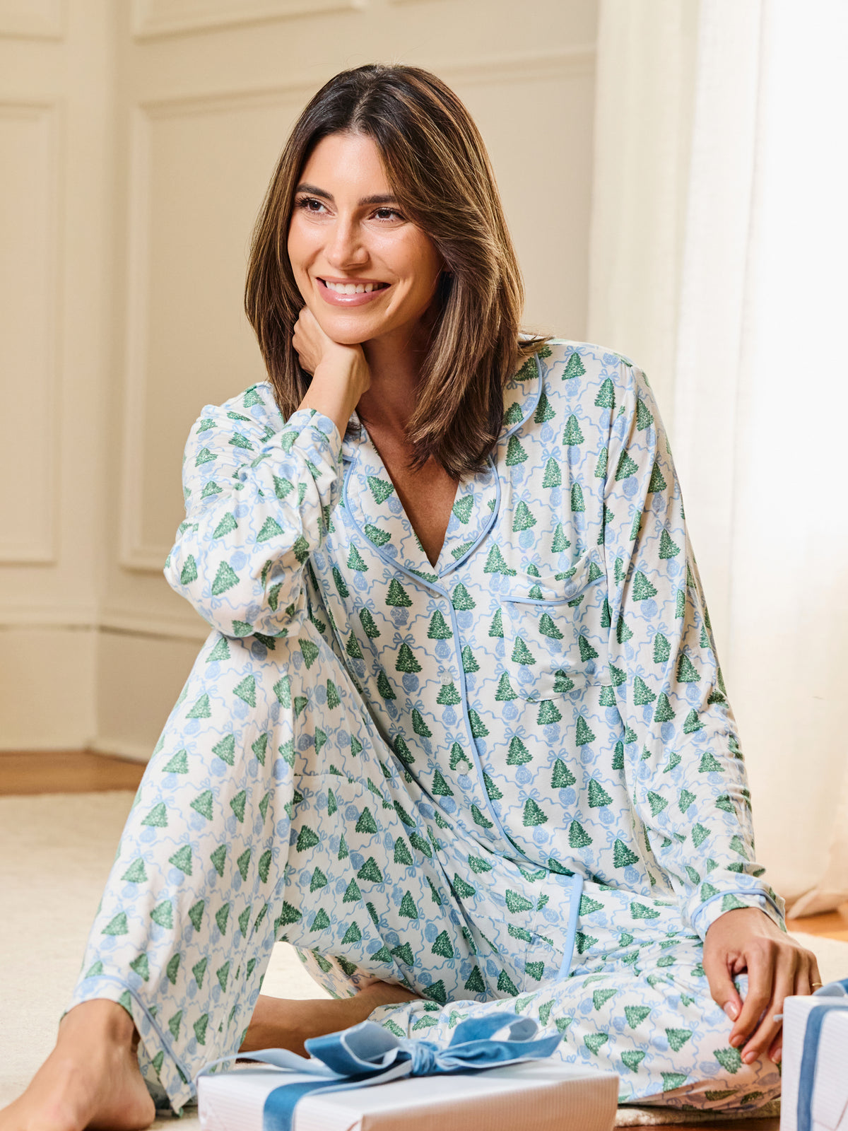 A woman wearing a light blue and white pajama set with a green pine tree pattern, sitting on a floor in a bright room with wrapped gifts in the foreground.