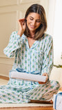 A woman sitting on a floor in a cozy room, wearing a light blue pajama set with a green pine tree pattern. She is holding a wrapped gift box with a blue ribbon, with a Christmas tree visible in the background.