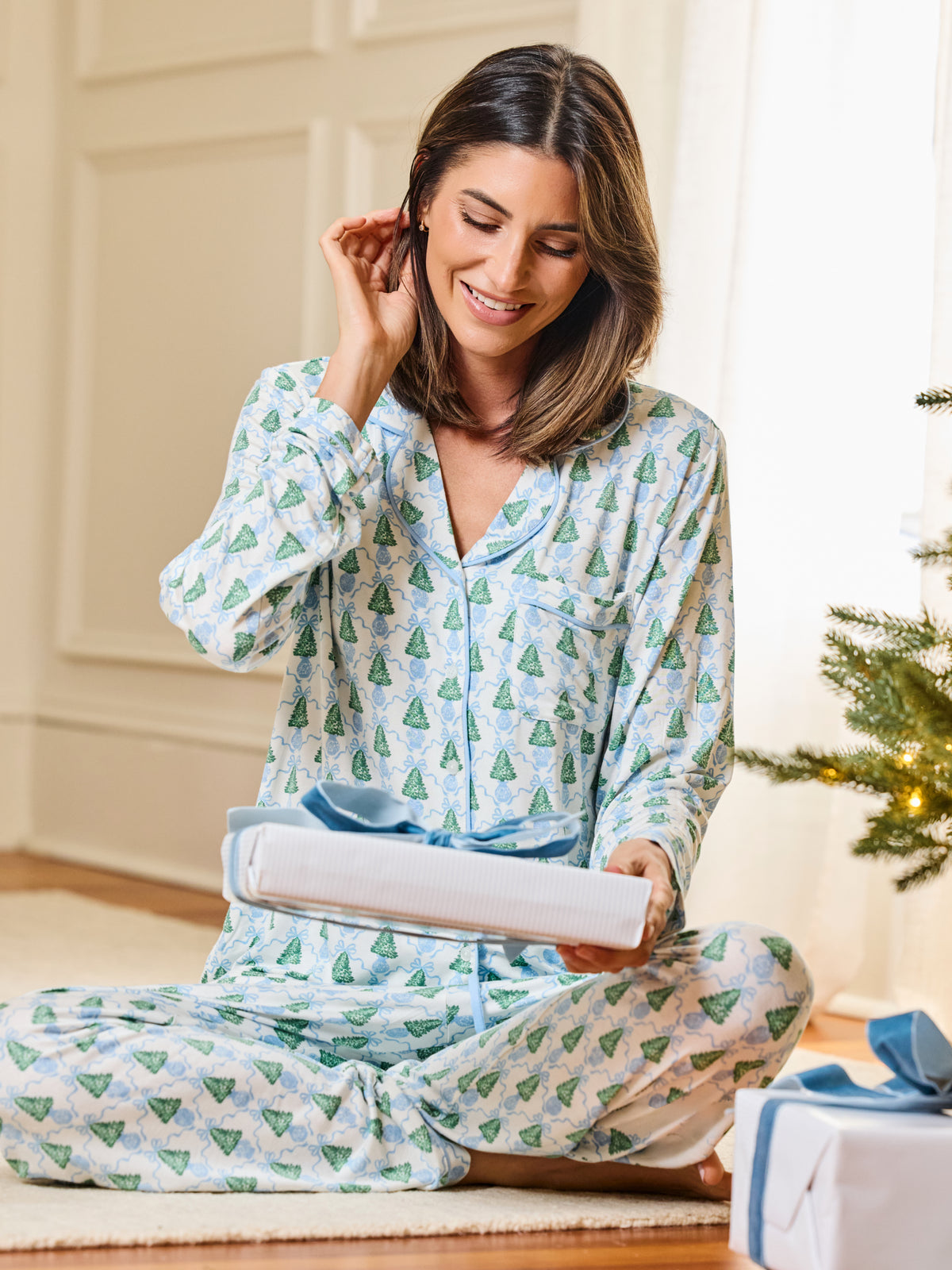 A woman sitting on a floor in a cozy room, wearing a light blue pajama set with a green pine tree pattern. She is holding a wrapped gift box with a blue ribbon, with a Christmas tree visible in the background.