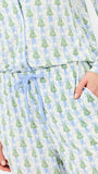 A close-up of a person wearing a long-sleeve pajama top and matching pants set with an all-over print of green Christmas trees and light blue bows on a white background. The set features a light blue drawstring belt and side pockets.
