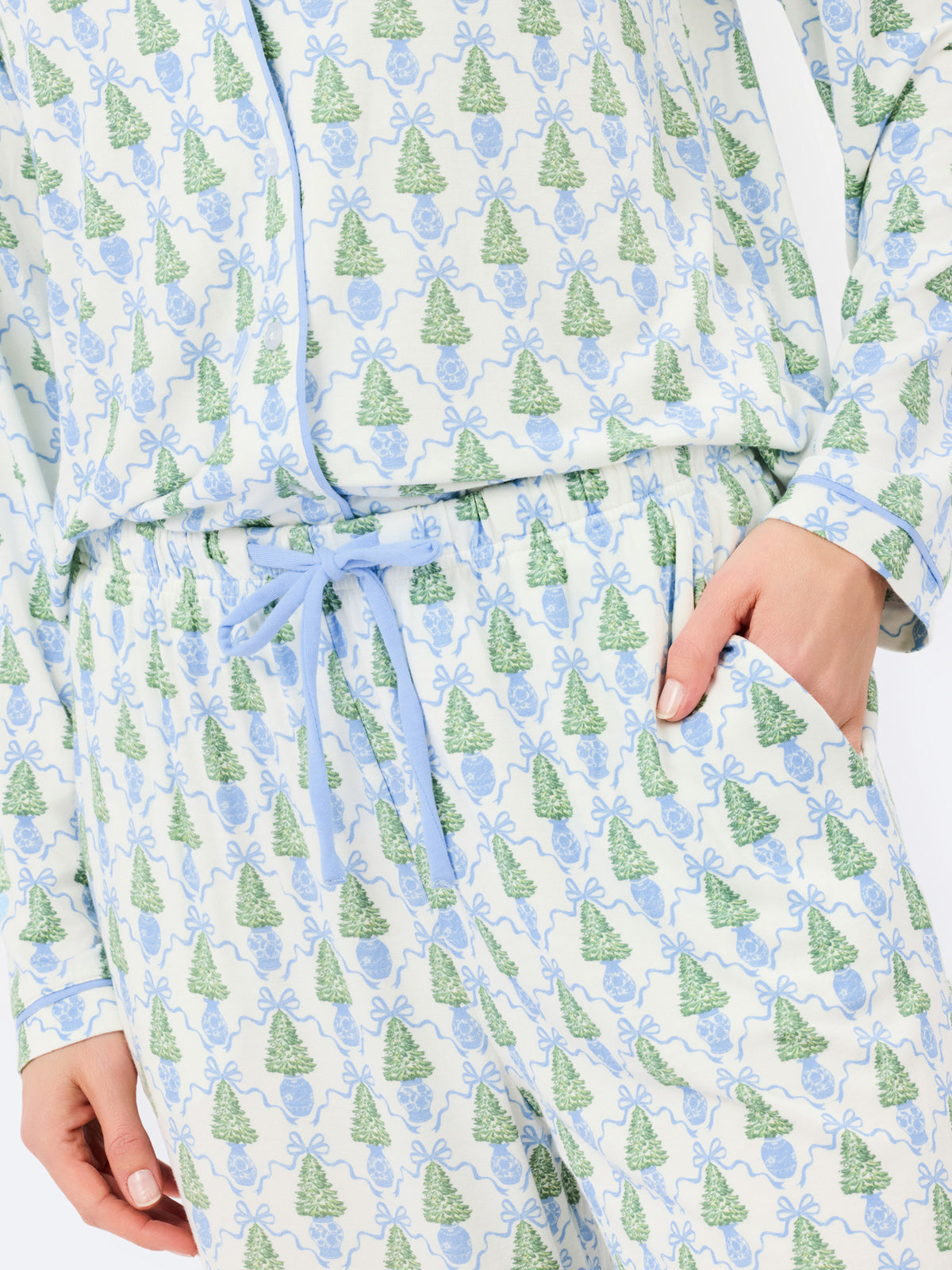 A close-up of a person wearing a long-sleeve pajama top and matching pants set with an all-over print of green Christmas trees and light blue bows on a white background. The set features a light blue drawstring belt and side pockets.