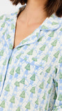 A close-up of a person wearing a pajama top with a white base, light blue piping, and an allover print of green Christmas trees and blue ornaments.