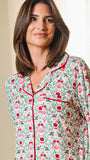 A woman wearing a long-sleeve pajama top with a festive Christmas-themed print of red roses, green leaves, and pink ornaments, featuring red piping on a light cream background.