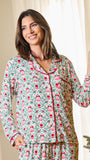 A woman wearing a festive Christmas-themed pajama set with a green background, red trim, and a pattern of red berries and pink ornaments, standing next to a Christmas tree.