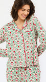 A woman wearing a long-sleeve pajama top and matching pants set with a festive green, red, and pink Christmas tree pattern on a cream background, set against a plain light gray studio background.
