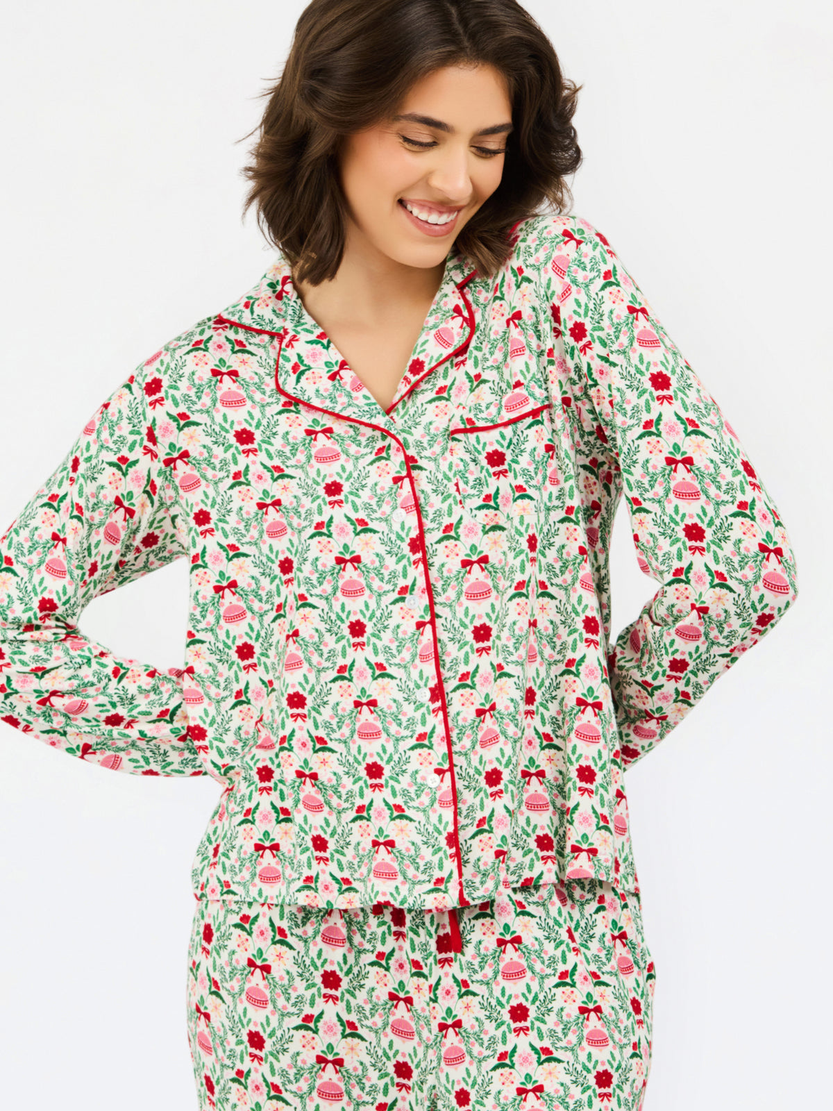 A woman wearing a long-sleeve pajama top and matching pants set with a festive green, red, and pink Christmas tree pattern on a cream background, set against a plain light gray studio background.