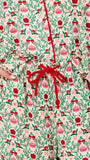 A close-up of a person wearing a long-sleeve holiday-themed jumpsuit or romper with a festive print of red bells, red roses, green leaves, and pink bows on a cream background, featuring a red drawstring waist.