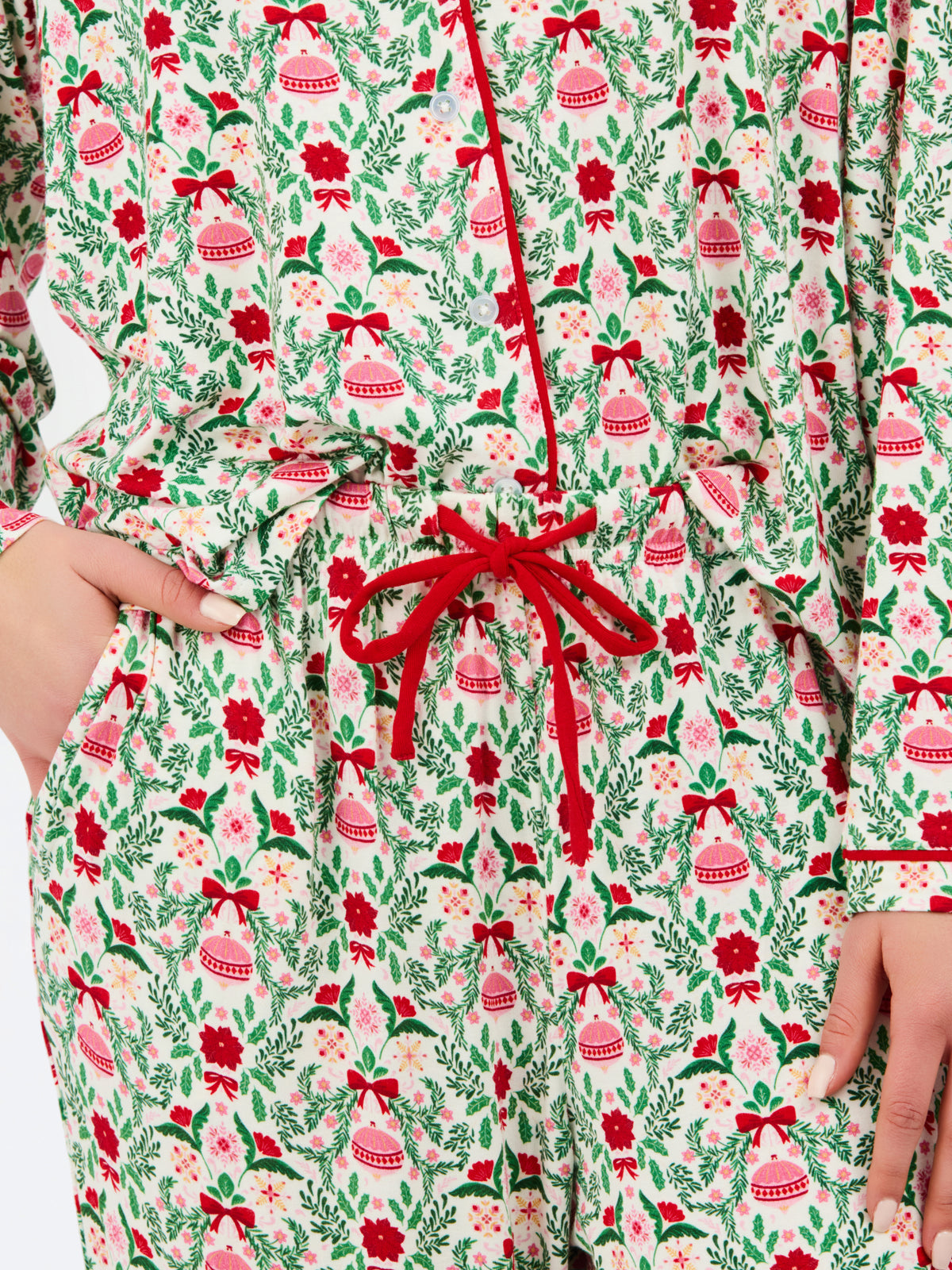 A close-up of a person wearing a long-sleeve holiday-themed jumpsuit or romper with a festive print of red bells, red roses, green leaves, and pink bows on a cream background, featuring a red drawstring waist.