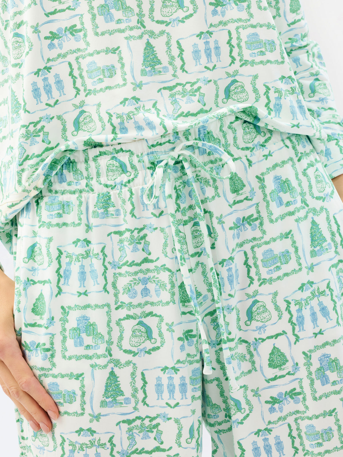 A close-up of a person wearing a matching pajama set with a festive holiday print. The set features a white base fabric decorated with green and light blue illustrations of Christmas trees, stockings, and gift boxes.