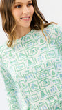 A woman wearing a long-sleeve crewneck top with a green and blue winter-themed pattern of snowflakes, trees, and houses on a white background.