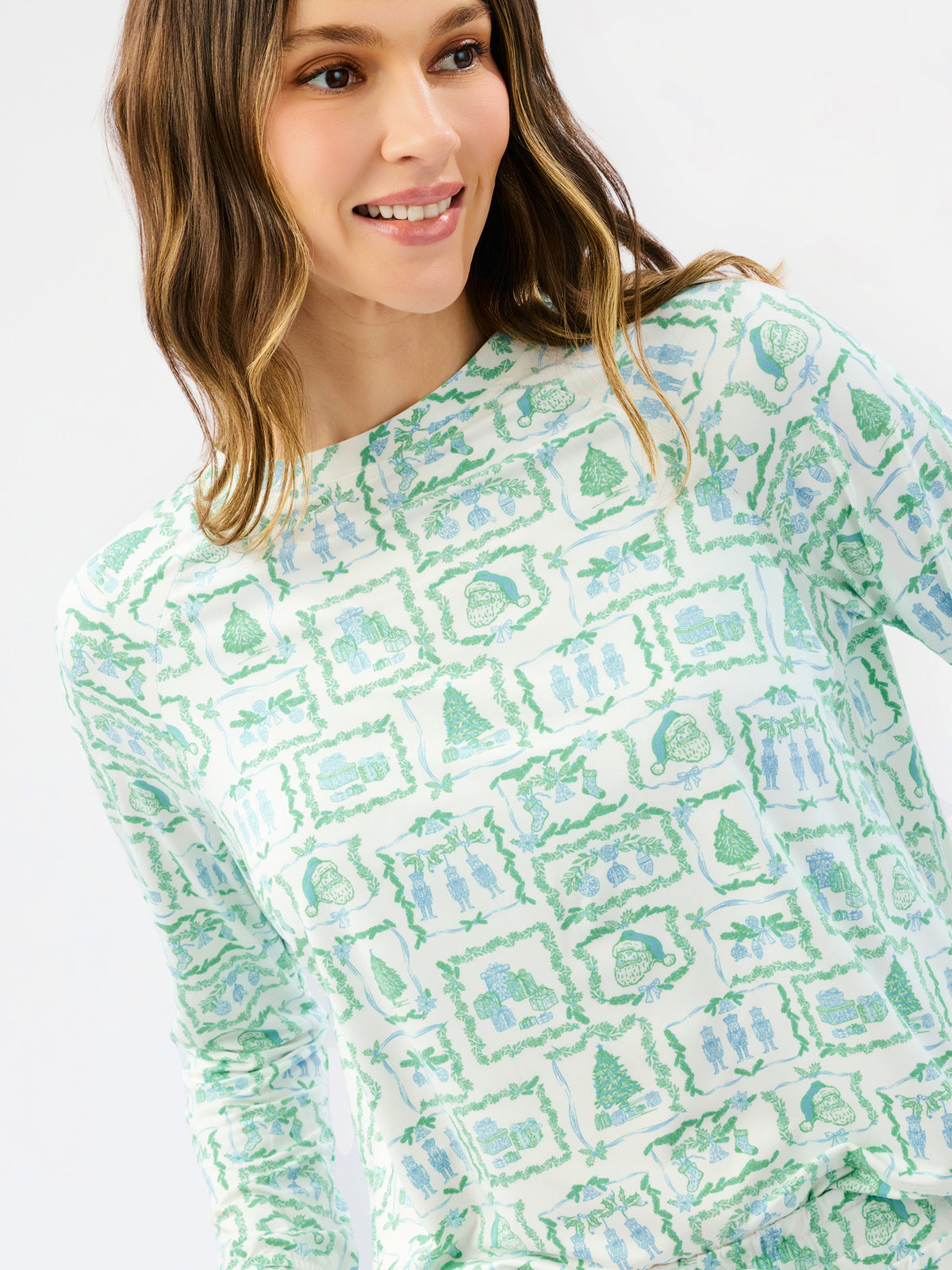 A woman wearing a long-sleeve crewneck top with a green and blue winter-themed pattern of snowflakes, trees, and houses on a white background.