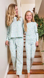 Two girls wearing matching long-sleeve pajamas with a light mint green and white geometric pattern, walking down a wooden staircase indoors next to a decorated Christmas tree.