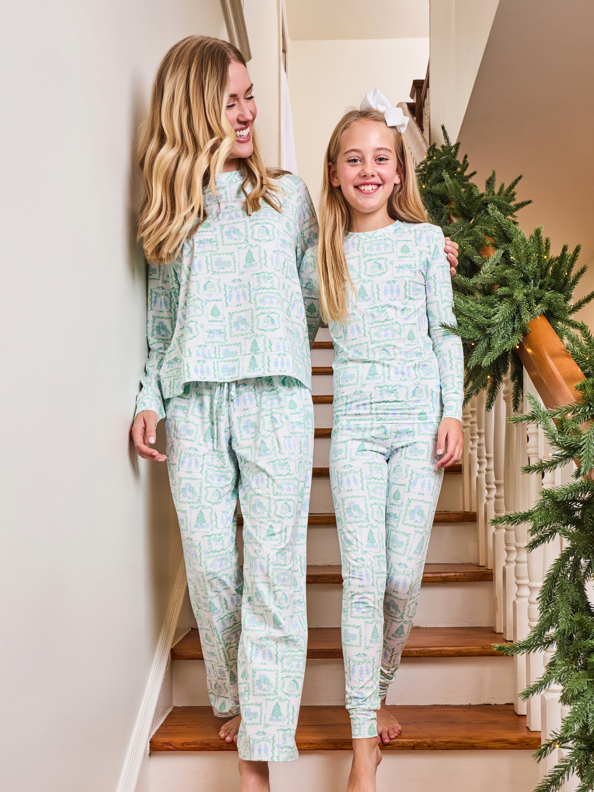 Two girls wearing matching long-sleeve pajamas with a light mint green and white geometric pattern, walking down a wooden staircase indoors next to a decorated Christmas tree.