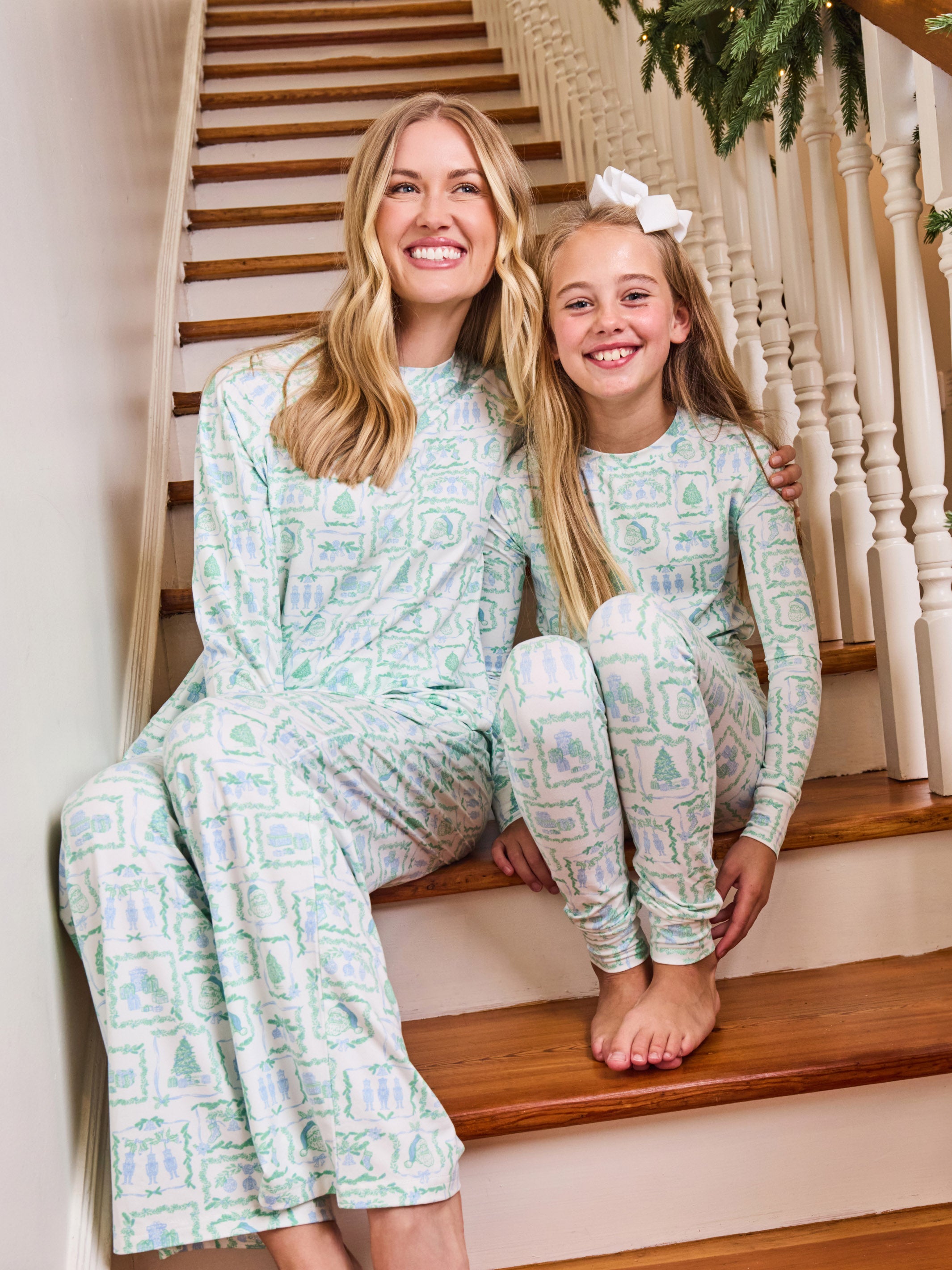 A woman and a young girl sitting together on wooden stairs indoors, wearing matching long-sleeve pajamas with a pale mint green and white ornamental pattern.