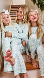 Three women of different ages wearing matching light green and white Christmas-themed patterned pajamas, sitting together on wooden stairs in a home setting.