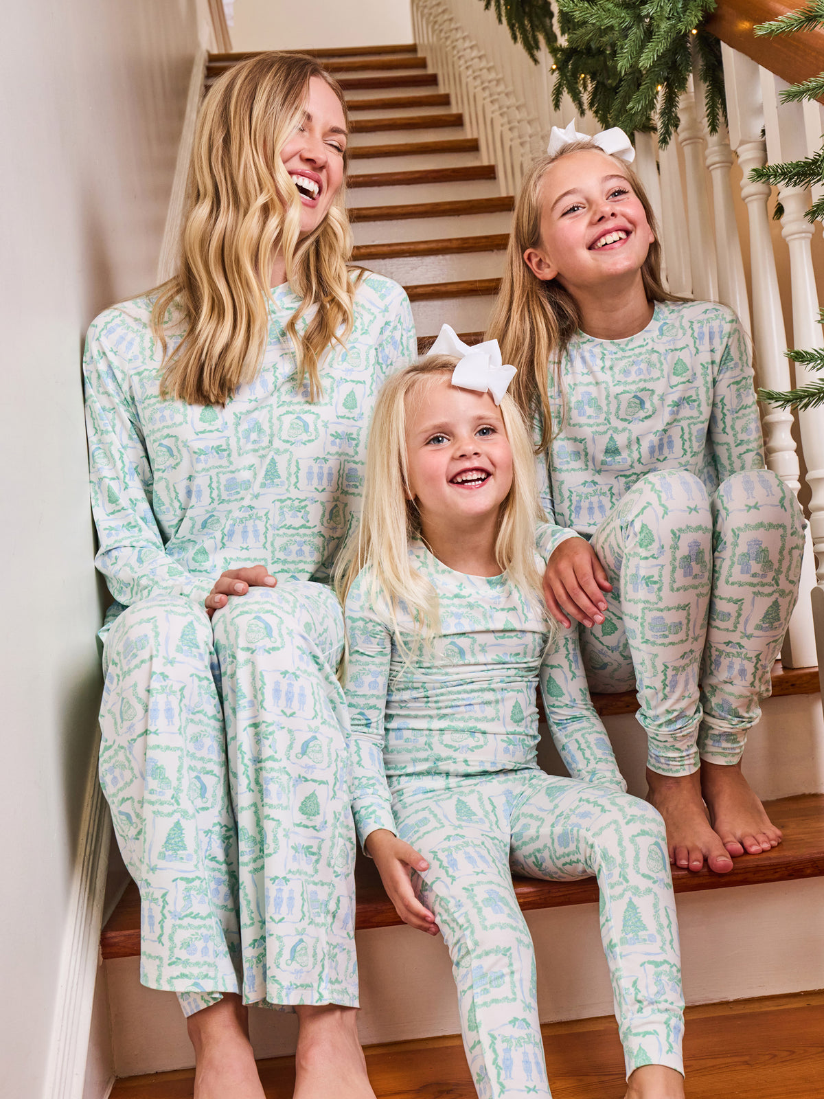 Three women of different ages wearing matching long-sleeve pajamas with a light green and white Christmas tree pattern, sitting together on wooden stairs in a holiday setting.