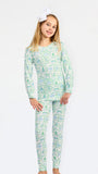 A young girl modeling a matching long-sleeve pajama top and pants set with a light green and white campfire pattern, featuring a white bow in her hair, set against a plain white studio background.