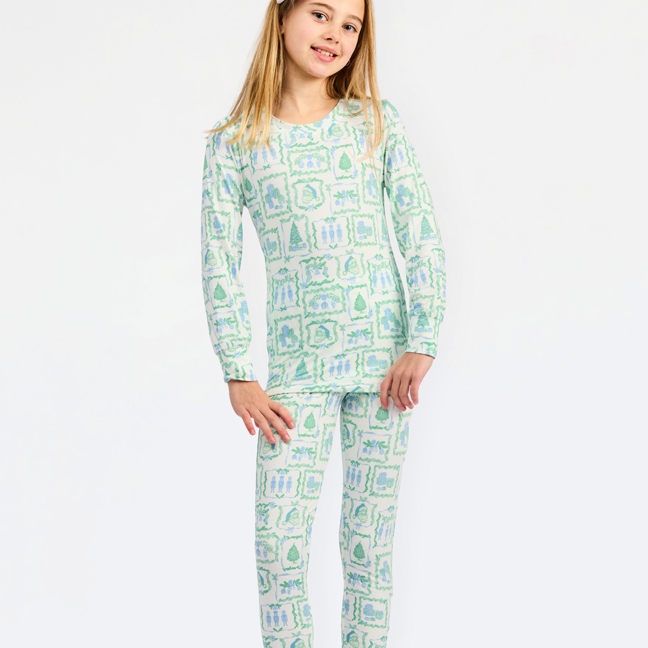 A young girl modeling a matching long-sleeve pajama top and pants set with a light green and white campfire pattern, featuring a white bow in her hair, set against a plain white studio background.