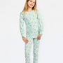 A young girl modeling a matching long-sleeve pajama top and pants set with a light green and white campfire pattern, featuring a white bow in her hair, set against a plain white studio background.