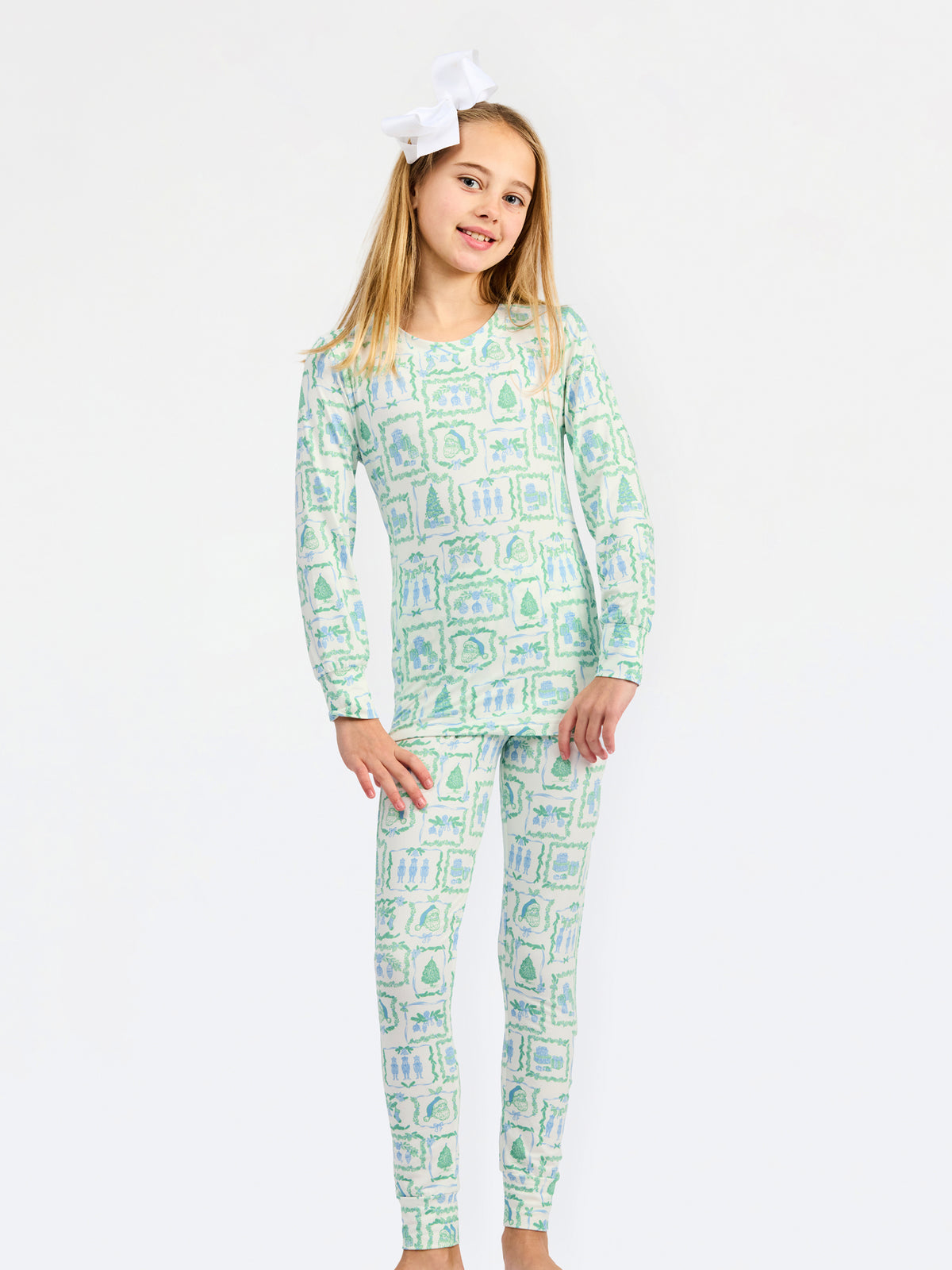 A young girl modeling a matching long-sleeve pajama top and pants set with a light green and white campfire pattern, featuring a white bow in her hair, set against a plain white studio background.
