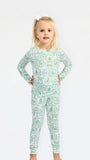 A young girl with blonde hair tied with a white bow stands against a plain white background wearing a long-sleeve one-piece pajama with a green and blue winter landscape pattern.