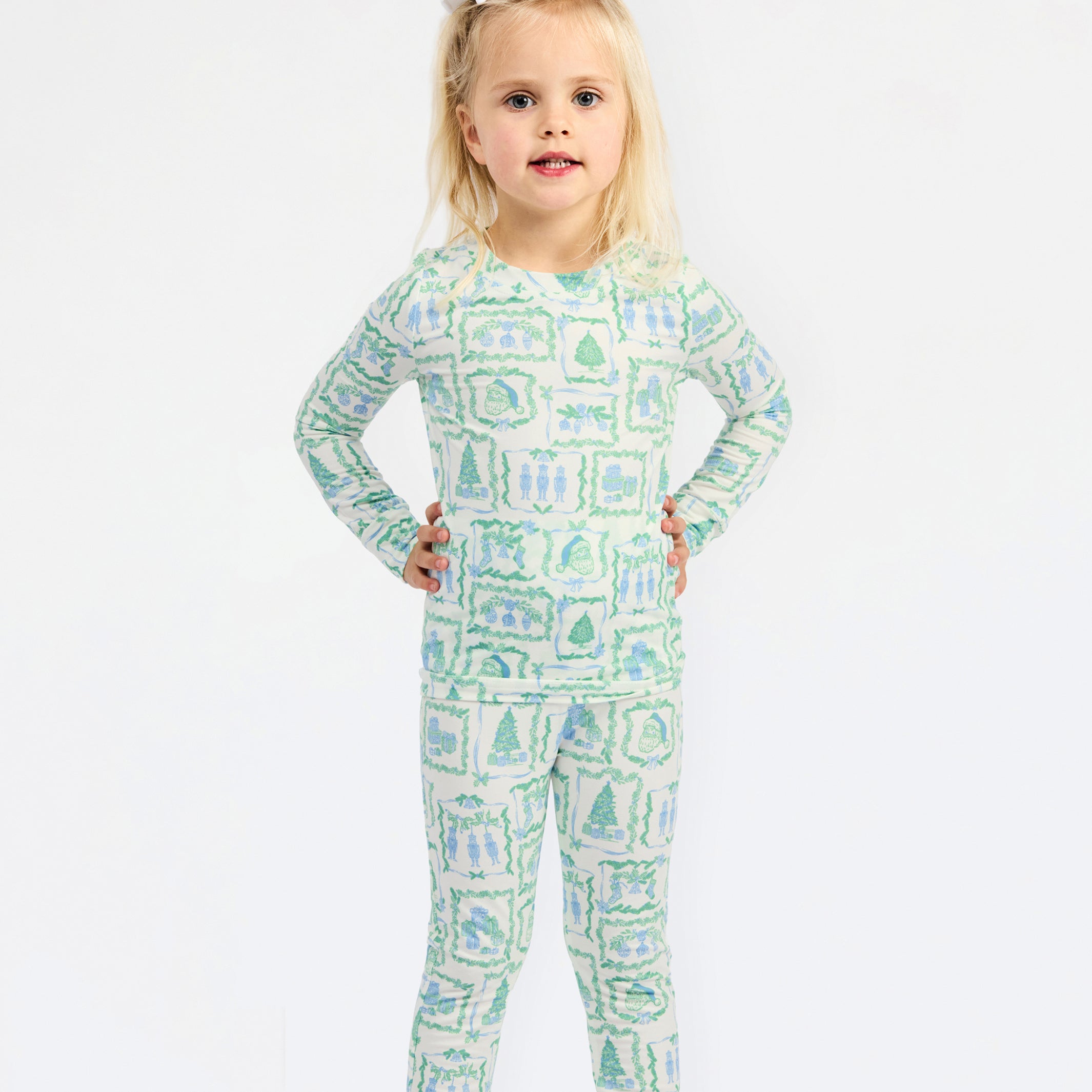 A young girl with blonde hair tied with a white bow stands against a plain white background wearing a long-sleeve one-piece pajama with a green and blue winter landscape pattern.