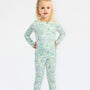 A young girl with blonde hair tied with a white bow stands against a plain white background wearing a long-sleeve one-piece pajama with a green and blue winter landscape pattern.