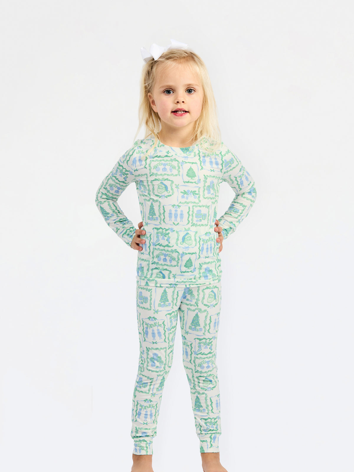 A young girl with blonde hair tied with a white bow stands against a plain white background wearing a long-sleeve one-piece pajama with a green and blue winter landscape pattern.