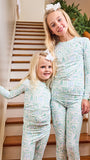 Two young girls wearing matching long-sleeve pajamas with a light blue and white Christmas tree pattern, standing on a wooden staircase indoors.