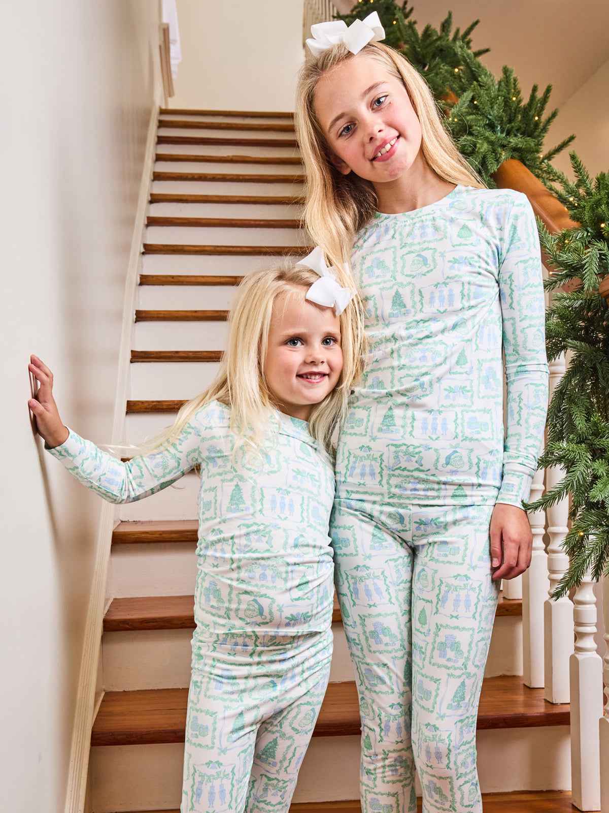 Two young girls wearing matching long-sleeve pajamas with a light blue and white Christmas tree pattern, standing on a wooden staircase indoors.