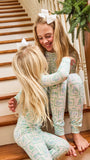 Two young girls sitting on wooden stairs indoors, wearing matching long-sleeve pajamas with a light mint green and white holiday village print. One girl has a white bow in her hair and is smiling at the other.