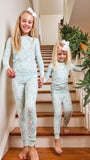 Two young girls walking down wooden stairs indoors, wearing matching long-sleeve and leggings pajama sets with a light mint green and white geometric pattern. The older girl wears a white bow in her hair, and the stairs are decorated with a green garland.